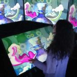 The image shows a person is interacting with a digital display inside an immersive exhibit at MADS Art Gallery. The screen depicts a vibrant and colourful digital art, depicting a stylized perfume bottle with a figure inside it. The bottle is primarily in hues of purple and blue, with an ethereal quality emphasized by surrounding swirling clouds and vapour-like elements in white and pink.. Multiple layers of the screen surrounding the person show repeating images of the same stylized perfume bottle shown on the main screen. The person appears to be touching or gesturing toward the display, engaged in an interactive component of the exhibit.