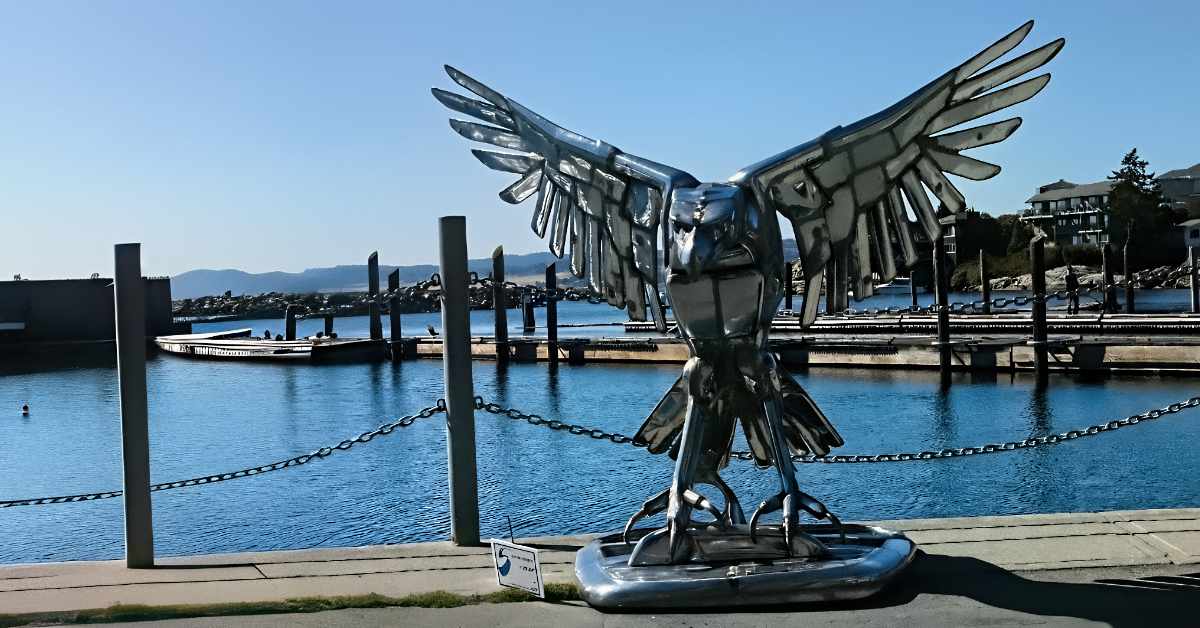 A metallic sculpture of an eagle stands prominently in the foreground, with its wings spread wide as if about to take flight. The sculpture's shiny surface reflects light, highlighting its detailed feathers and powerful stance. Behind the sculpture, a calm blue harbor with several docks extends into the distance. The background features gently rolling hills under a clear sky, with a smattering of buildings at the water's edge. A single chain-link barrier runs along the waterfront.