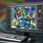 The image shows a desktop computer setup, including a monitor, keyboard, mouse, and a tower case. The monitor displays a digital art software with a colorful abstract artwork in progress, featuring swirling lines, brushstrokes in blue, green, yellow, and black, and a square frame in the center containing ornate black patterns. The user interface of the software includes various toolbars on the sides, with color options and drawing tools visible. The computer tower is silver and black, and the background features a soft, blurred, multicolored pattern.