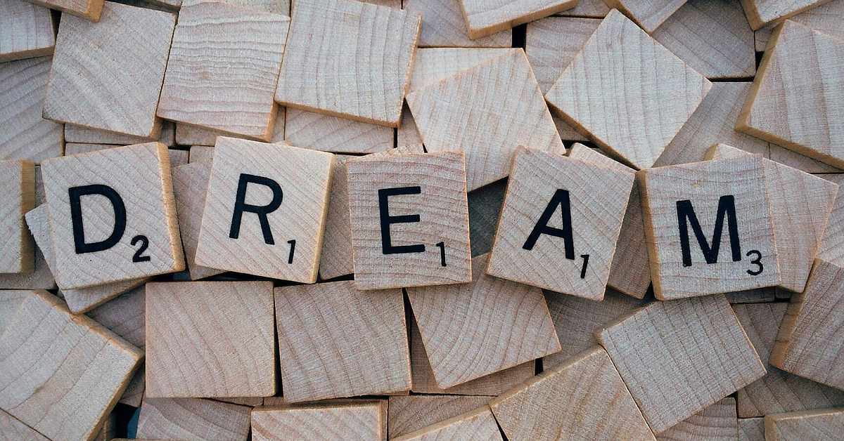 The image shows wooden Scrabble-style tiles scattered in a pile. Five tiles in the center spell out the word "DREAM" in black, uppercase letters. Each tile has a small number in the lower right corner indicating its point value, such as "D" worth 2 points and "M" worth 3 points. The surrounding tiles are blank or face down, creating a textured wooden background.
