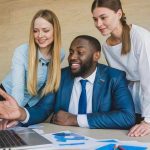 A group of three colleagues gathered around a laptop in a modern office setting. A man in a blue suit and tie is seated, smiling, and gesturing towards the laptop screen while speaking. Two women, one in a light blue shirt and the other in a white blouse, stand on either side of him, leaning in and looking at the screen attentively. Papers with charts and graphs are scattered on the desk in front of them. The background features a light wooden wall.