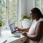 A woman wearing glasses and a sleeveless top sits at a white desk near a large window, working on a laptop. She is pointing at a chart displayed on the screen. A calculator, papers, and a notebook are on the desk. The space is decorated with potted plants, including a trailing vine and a large leafy plant in white ceramic pots. The window allows natural light to enter, with trees visible outside.