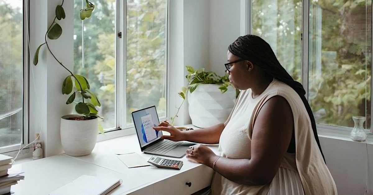 A woman wearing glasses and a sleeveless top sits at a white desk near a large window, working on a laptop. She is pointing at a chart displayed on the screen. A calculator, papers, and a notebook are on the desk. The space is decorated with potted plants, including a trailing vine and a large leafy plant in white ceramic pots. The window allows natural light to enter, with trees visible outside.