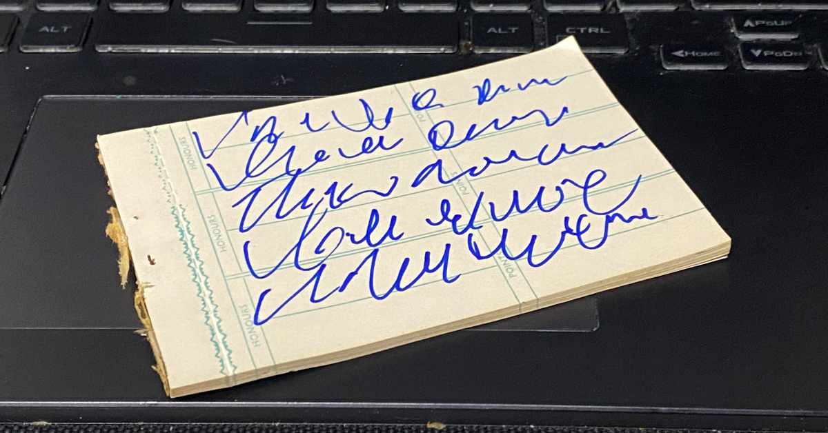The image shows a small notepad with rough edges, lying on a black computer keyboard. The notepad has faint blue grid lines and columns labeled with text, and it contains several lines of blue cursive handwriting that are highly scribbled and illegible. The writing appears to be hurried or shorthand, making the words indistinguishable.