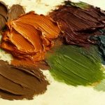 A close-up photo of thick, textured blobs of oil paint in earthy and autumn-inspired tones arranged on a light-colored palette. The colors closely resemble the October 2025 palette, including dark brown, burnt orange, burgundy, beige, green, and blue-gray hues, with visible brush strokes adding texture and depth.